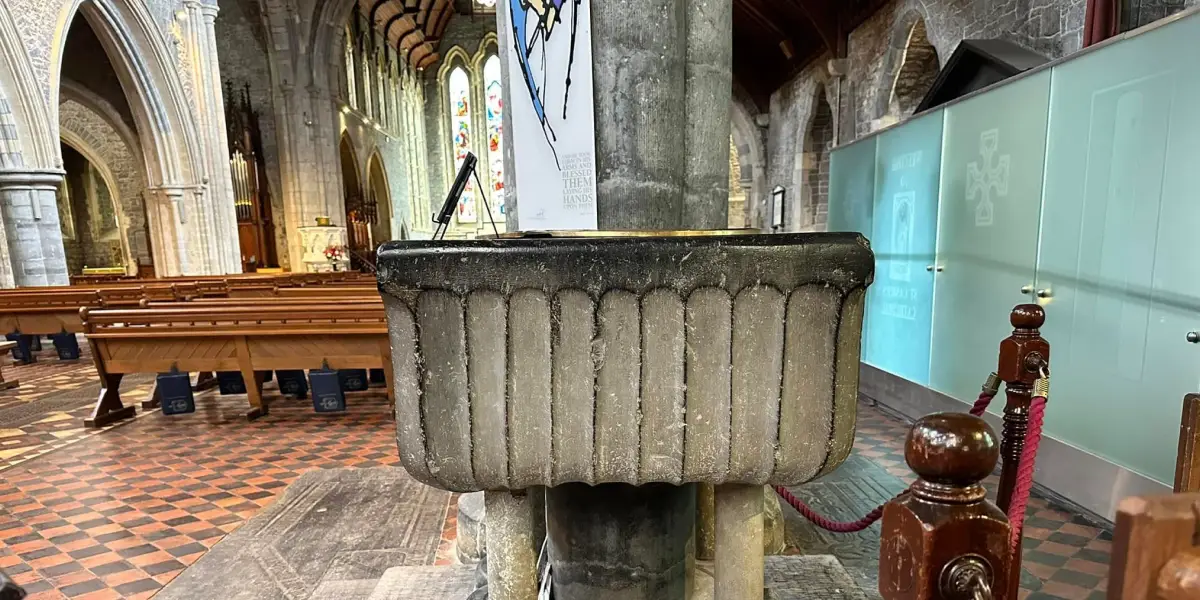 A stone baptismal font inside a historic church with high arched ceilings. Wooden pews and stained glass windows are visible in the background, creating a serene atmosphere. A rope barrier surrounds the font.