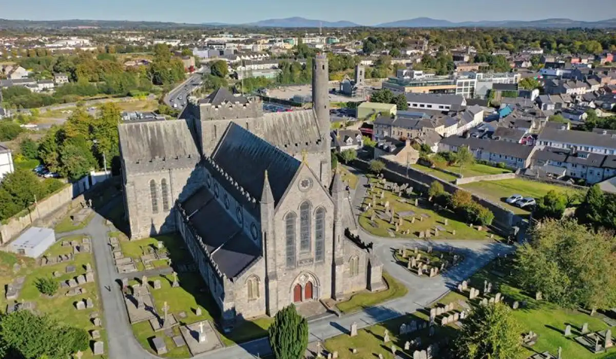 Aerial view of a large stone church with a tall spire surrounded by a graveyard and greenery. The church is situated in a town with nearby houses, buildings, and distant hills under a clear blue sky.