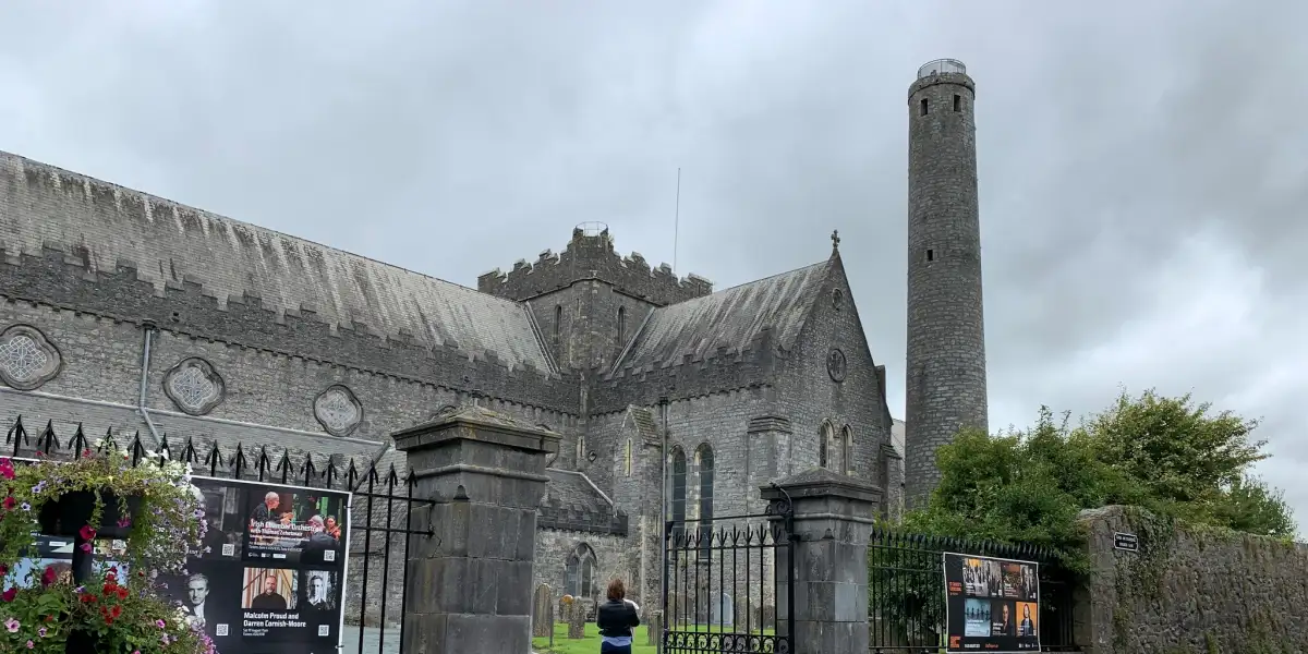 A historic stone cathedral with a tall, cylindrical tower stands amid an old graveyard with weathered tombstones. The sky is clear and blue, and a tree with green leaves partially frames the right side of the image.