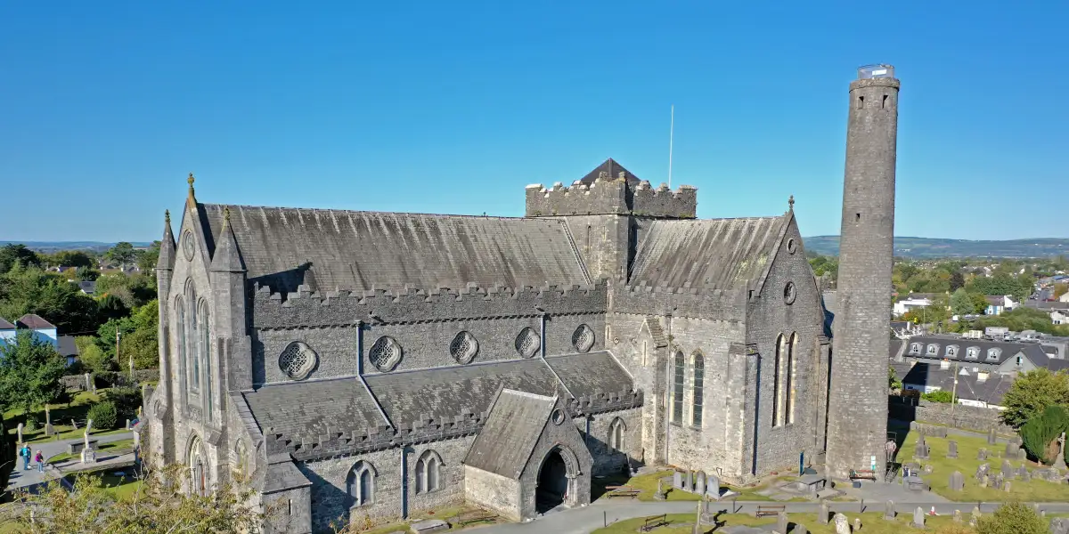 A historic stone cathedral with a tall, cylindrical tower stands amid an old graveyard with weathered tombstones. The sky is clear and blue, and a tree with green leaves partially frames the right side of the image.
