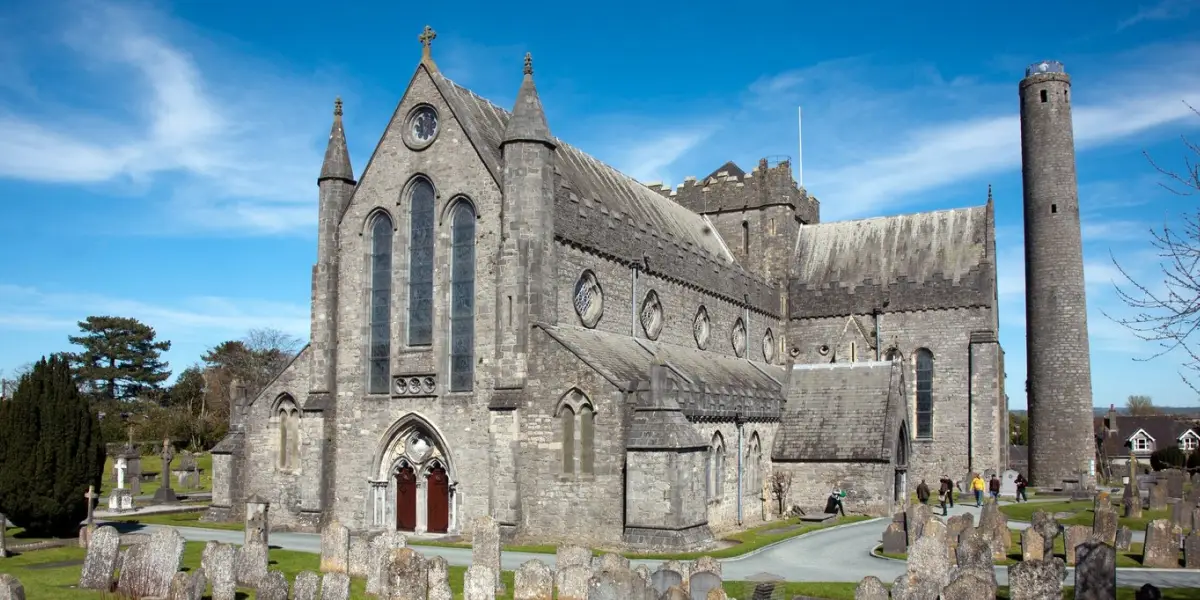 A historic stone cathedral and round tower set within a graveyard under a clear blue sky. The cathedral has tall, arched windows and intricate architectural details. Several gravestones are visible in the foreground.