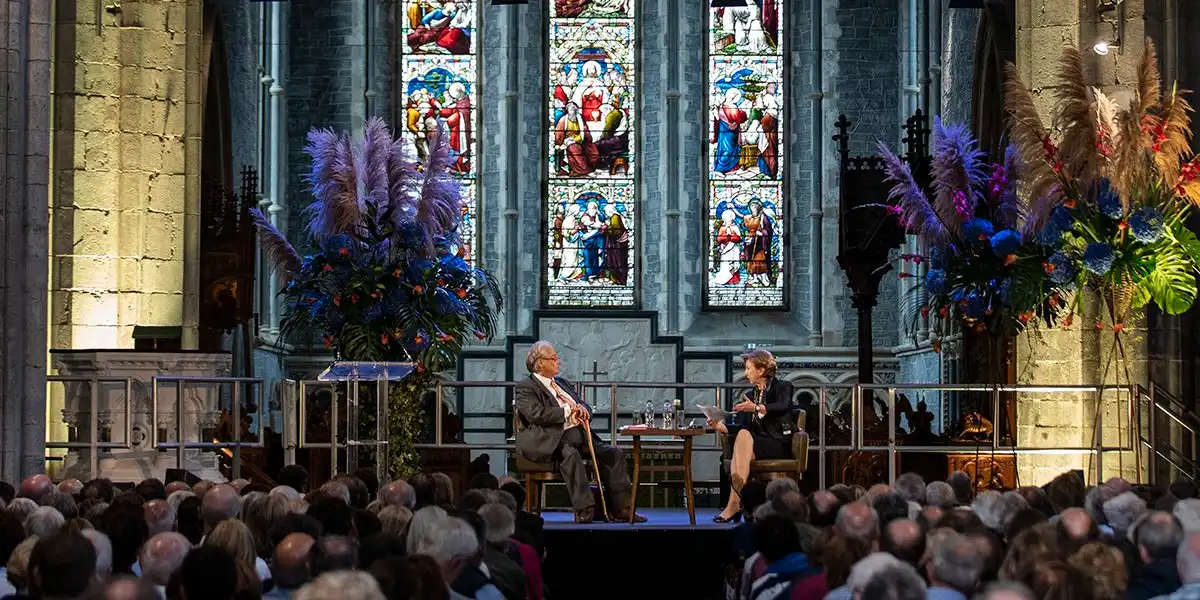 Two individuals are seated on a stage engaging in discussion inside a church with an audience watching. The backdrop features large stained glass windows and floral arrangements.